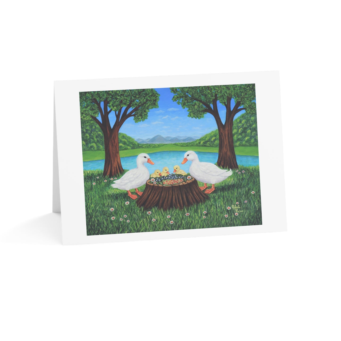 A folded white greeting card standing upright on a white background, printed with an illustration of two white ducks sitting on a tree stump in a grassy park by a lake, with trees, blue sky, and distant hills.