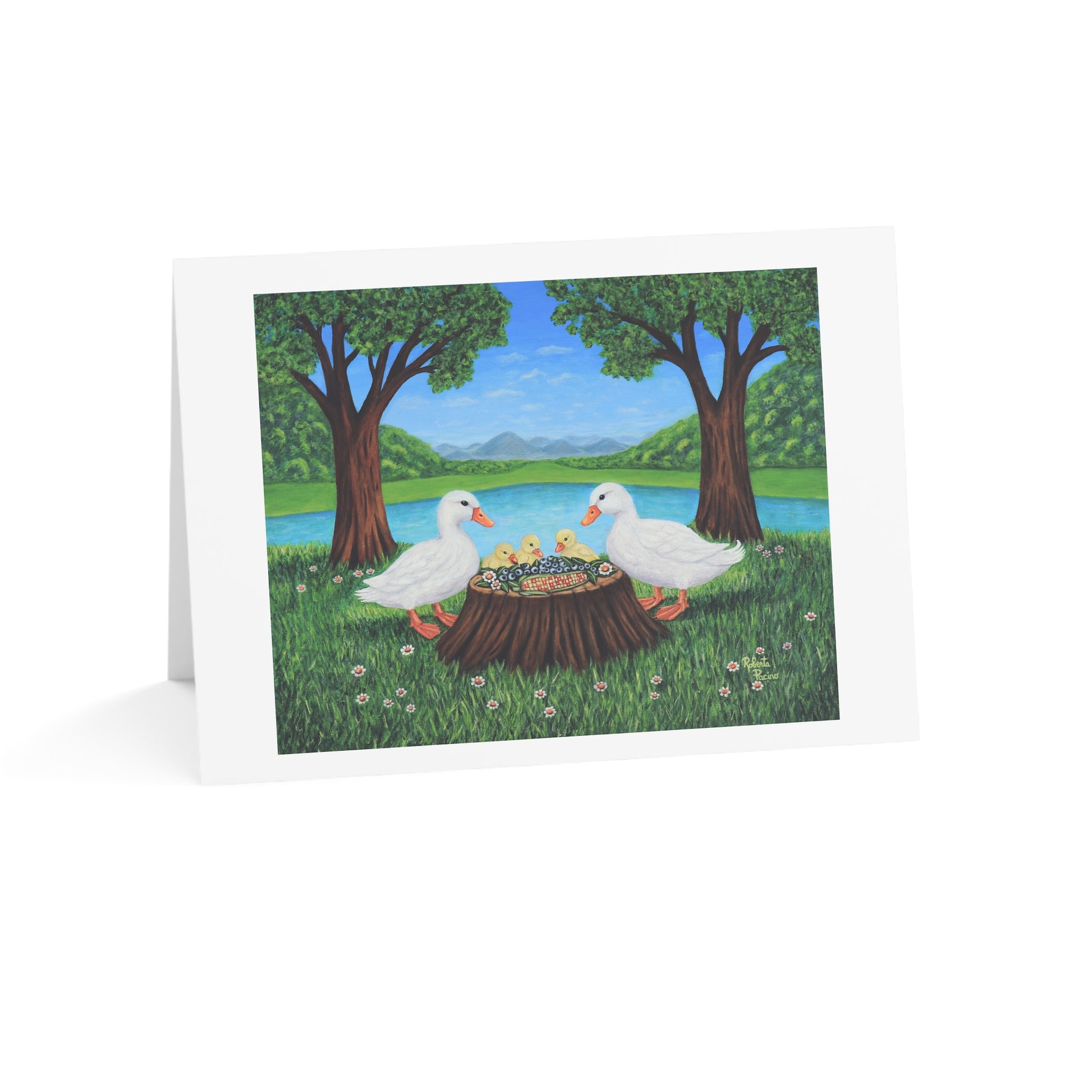 A folded white greeting card standing upright on a white background, printed with an illustration of two white ducks sitting on a tree stump in a grassy park by a lake, with trees, blue sky, and distant hills.