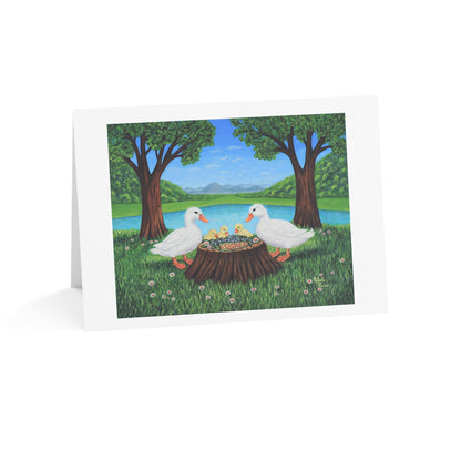 A folded white greeting card standing upright on a white background, printed with an illustration of two white ducks sitting on a tree stump in a grassy park by a lake, with trees, blue sky, and distant hills.