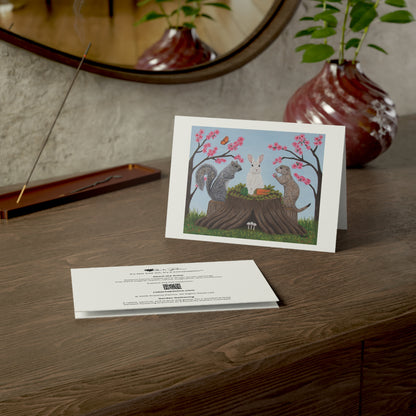 A printed art card or invitation with a whimsical illustration of a squirrel, prairie dot and a rabbit at a tree stump, displayed on a wooden table next to the back of another card, with a vase and mirror in the background.
