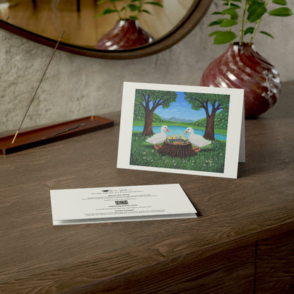 A printed art card or invitation standing on a wooden table, showing an illustration of two white geese at a tree stump by a lake, with the back of another card placed in front. A vase, mirror, and incense holder are visible in the styled setting.