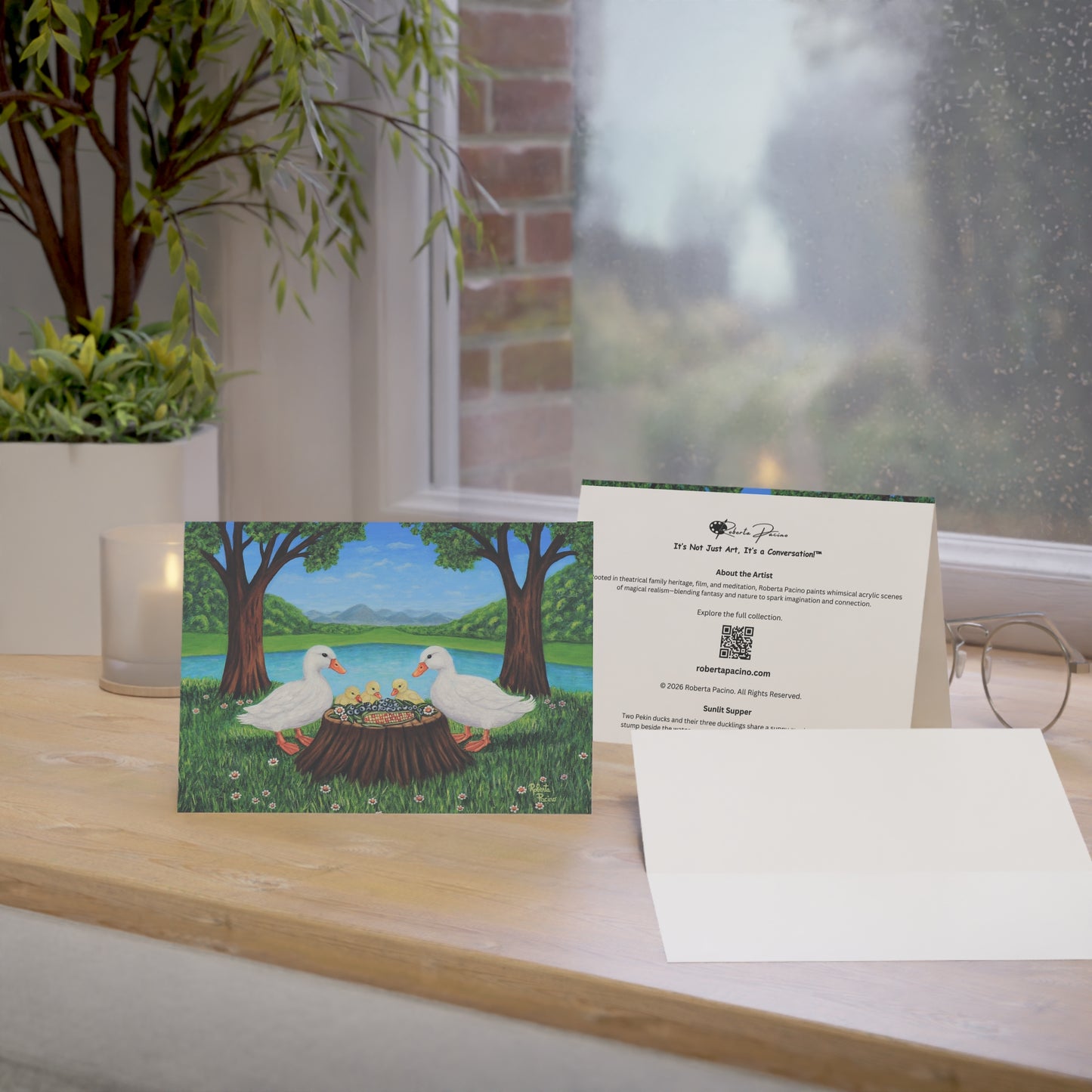 Sunlit Supper greeting card featuring two white ducks and three ducklings sharing a meal beside a pond in a sunny park setting, from original art by Roberta Pacino