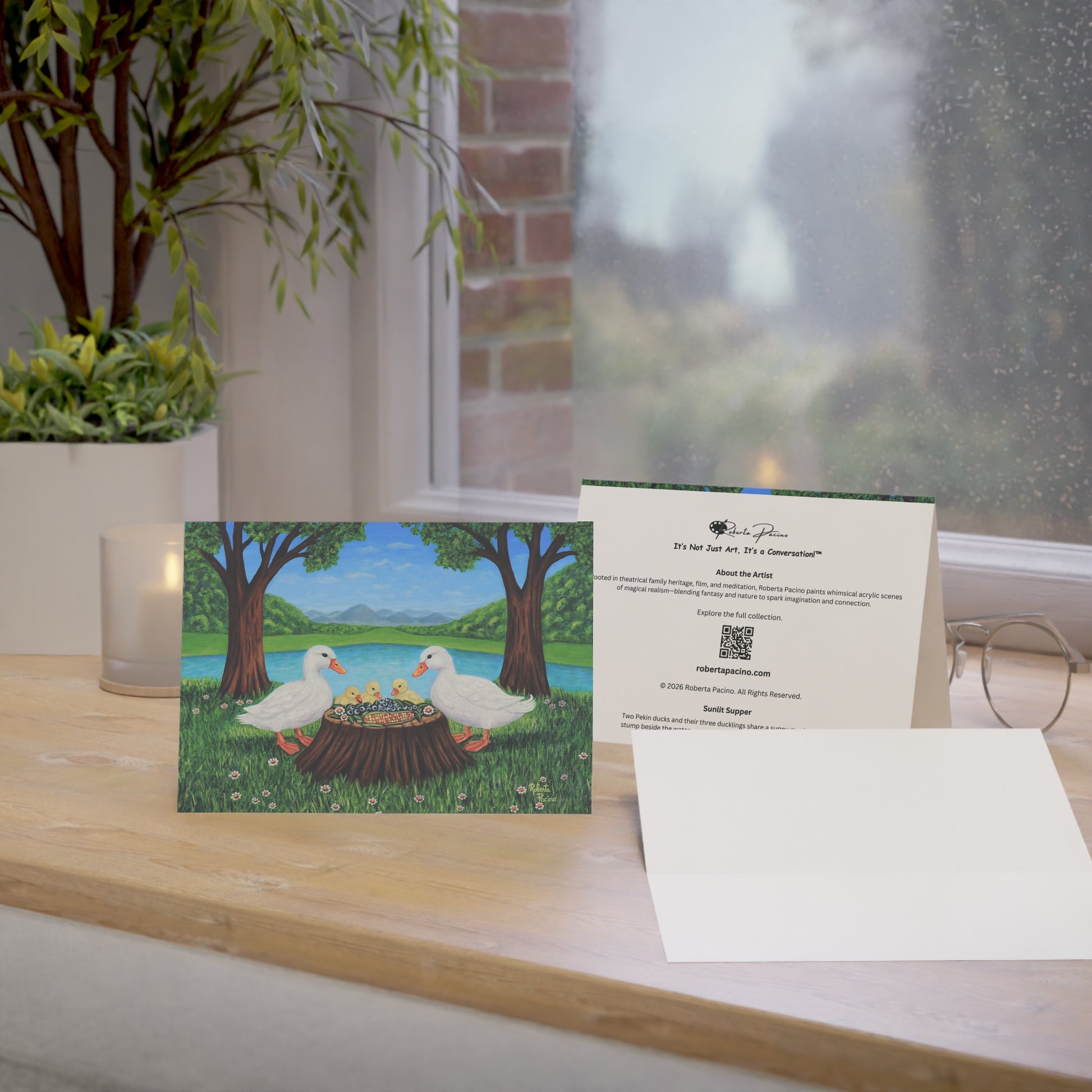 Sunlit Supper greeting card featuring two white ducks and three ducklings sharing a meal beside a pond in a sunny park setting, from original art by Roberta Pacino