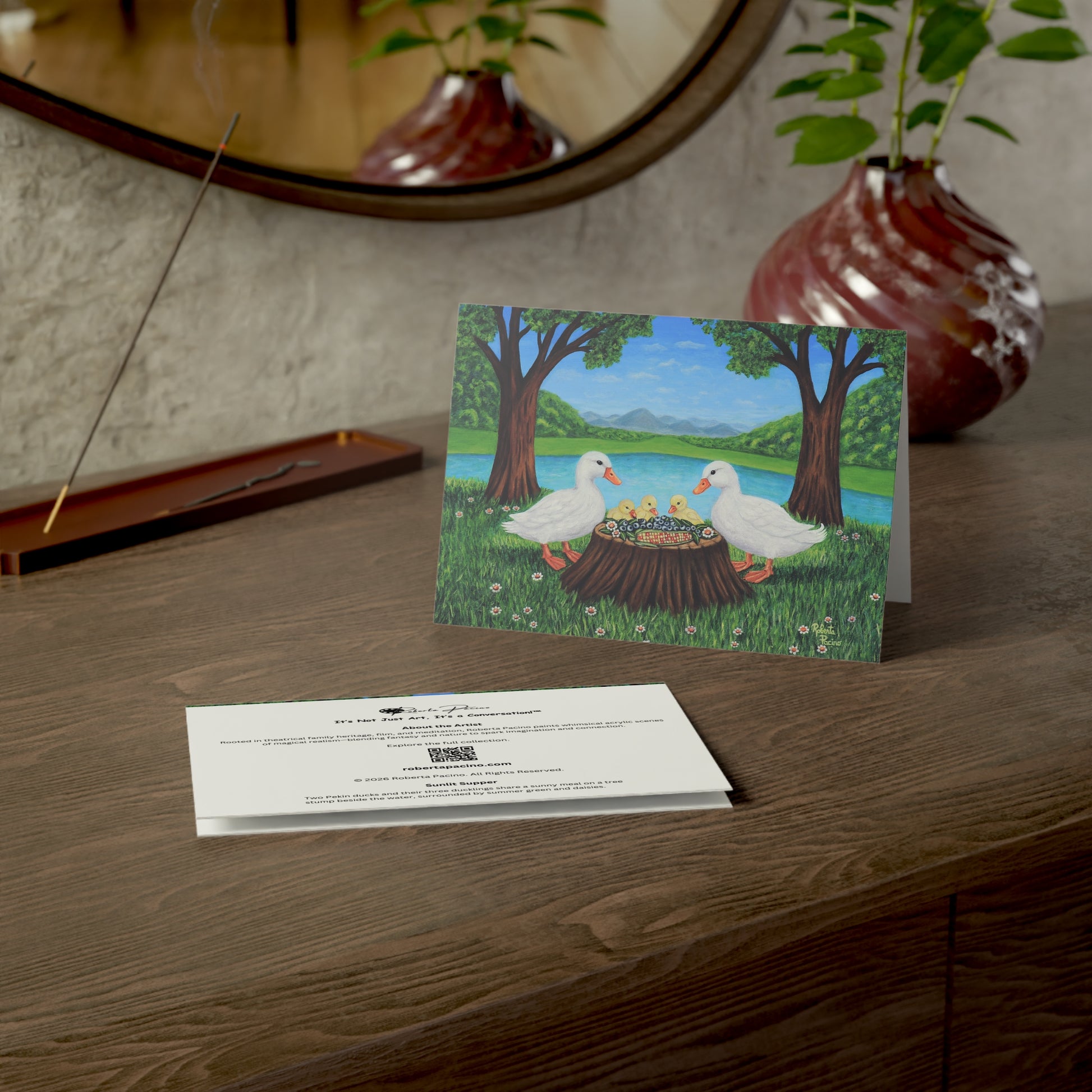 Sunlit Supper greeting card featuring two white ducks and three ducklings sharing a meal beside a pond in a sunny park setting, from original art by Roberta Pacino